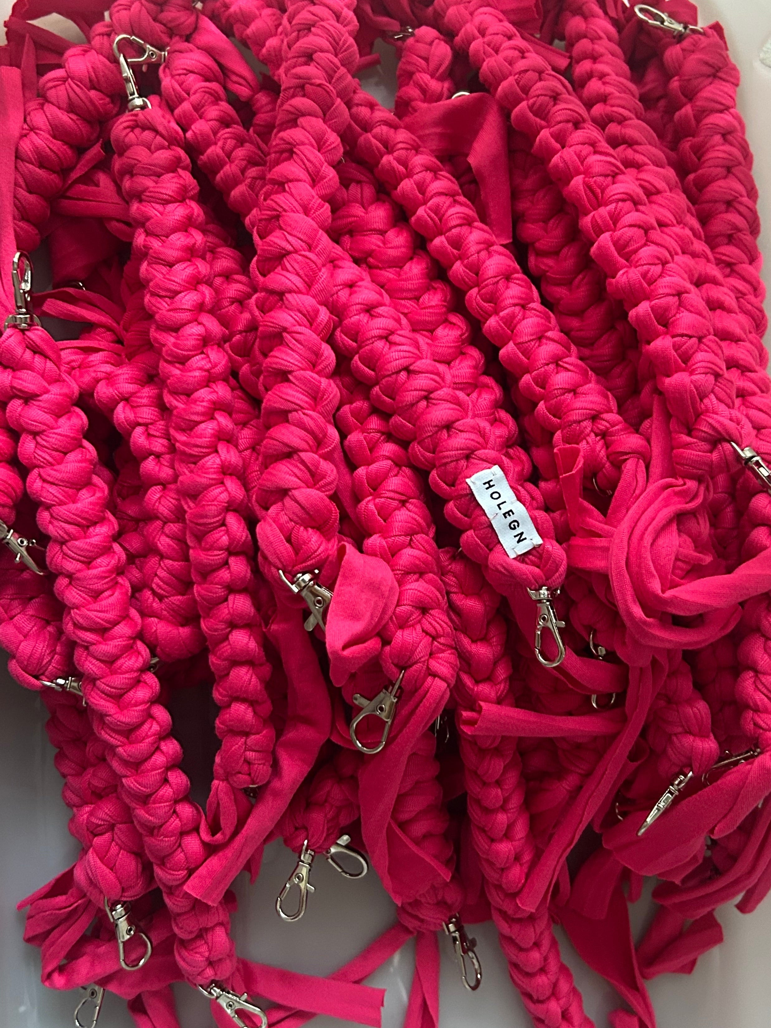 A pile of bright pink crochet phone straps with silver metal clasps and a small white tag reading 'HOLEGN' on one of the straps, photographed close-up on a light surface.